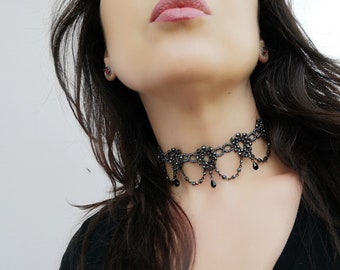 May include: A black choker necklace with a delicate lace design and dangling black beads. The necklace is worn by a person with long dark hair.