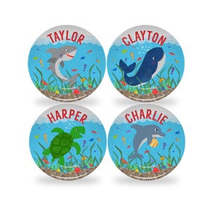 May include: Four personalized blue and white plates with a sea theme. Each plate features a different sea creature: a shark, a whale, a turtle, and a dolphin. The plates are decorated with seaweed, coral, and other sea life. The names "TAYLOR", "CLAYTON", "HARPER", and "CHARLIE" are printed in red on each plate.