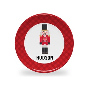 May include: Red and white personalized Christmas plate with a nutcracker illustration and the name "HUDSON" printed on the front.