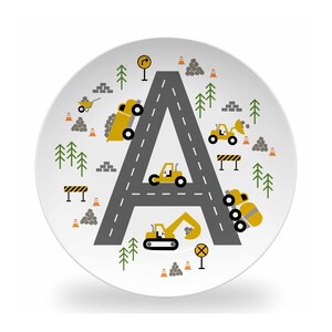 May include: A white ceramic plate with a gray road shaped like the letter A. The road is decorated with yellow construction vehicles, including a dump truck, a bulldozer, a backhoe, and a cement mixer. There are also green trees, orange traffic cones, and gray rocks on the plate.
