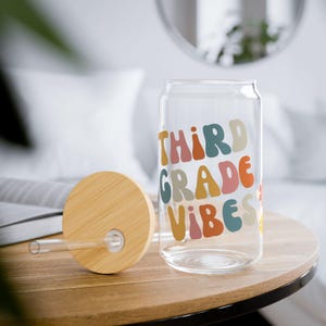 May include: A clear glass can-shaped tumbler with a wooden lid and straw. The tumbler features the text "THIRD GRADE VIBES" in colorful, retro-style lettering. The lid and straw are made of natural wood and clear plastic.