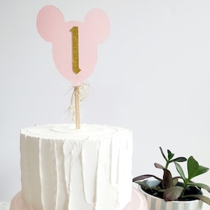 May include: A white cake with pink frosting on a pink cake stand. The cake is topped with a pink and gold Mickey Mouse head with the number 1 on it. A small potted plant is in the background.