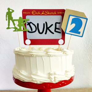Pode incluir: Um bolo branco com cobertura branca em um suporte de bolo vermelho. O bolo é decorado com um brinquedo Etch A Sketch vermelho e branco com o nome "DUKE" desenhado nele, um homenzinho verde do exército e um bloco azul com o número "2" nele.