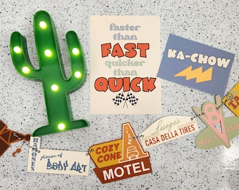 Puede incluir: Un conjunto de decoración de fiesta con temática de Cars con una luz de cactus verde, un cartel que dice "faster than FAST quicker than QUICK", un cartel que dice "KA-CHOW", un cartel que dice "TASTE-IN", y una cadena de carteles con el texto "TOW MATER", "RAMONES", "House of BODY ART", "COZY CONE MOTEL", "Luigi's CASA DELLA TIRES", "Flo's Cafe".