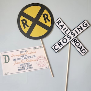 Puede incluir: Un conjunto de dos decoraciones de fiesta con temática de tren. Una es una señal redonda de cruce de ferrocarril amarilla y negra con las letras "RR" en negro. La otra es una señal blanca con letras negras que dice "RAILROAD CROSSING".