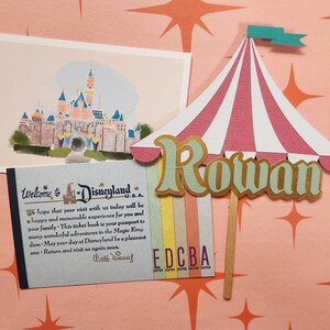 May include: A Disneyland-themed display featuring a postcard of a castle, a ticket book, and a circus tent-shaped sign with the name "Rowan." The sign has red and white stripes and a teal flag. The ticket book reads "Welcome to Disneyland U.S.A."