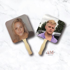 May include: Two square-shaped hand fans with wooden handles. The fans feature photos of two people, one woman and one man. The fans are on a white marble background.