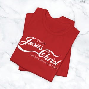 May include: A red t-shirt with white text that reads "Enjoy Jesus Christ" in a stylized font, with the phrase "And thou shalt never thirst" below. The shirt is folded on a white marble surface.
