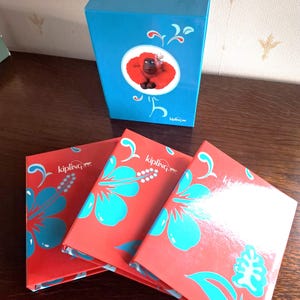 Könnte beinhalten: Eine leuchtend blaue rechteckige Schachtel mit einer kreisförmigen Grafik eines roten Orang-Utans, hinter drei roten rechteckigen Gegenständen, die mit großen türkisfarbenen Hibiskusblüten und dem weißen Kipling-Logo verziert sind.