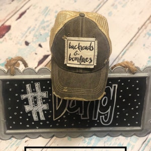 Puede incluir: Una gorra de camionero vintage negra con una malla dorada en la parte posterior. La gorra tiene un aspecto desgastado y presenta un parche con el texto "backroads & bonfires".