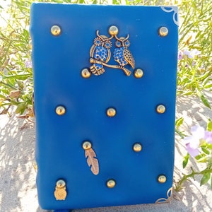 Puede incluir: Un diario de cuero azul con detalles dorados y un par de búhos en la portada. El diario tiene un borla y un diseño de pluma.