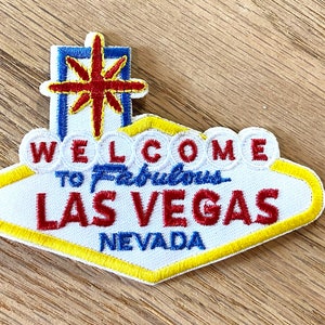 Puede incluir: Parche bordado blanco, amarillo y azul con el texto "Welcome to Fabulous Las Vegas Nevada". El parche presenta una estrella roja con un contorno azul.