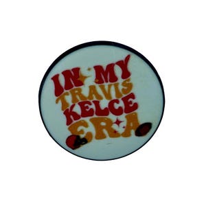 Puede incluir: Un bot&oacute;n redondo blanco con un borde negro. El bot&oacute;n tiene texto rojo y amarillo que dice "In My Travis Kelce Era".