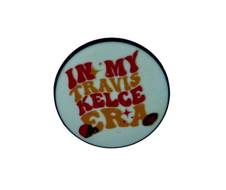 Puede incluir: Un bot&oacute;n redondo blanco con un borde negro. El bot&oacute;n tiene texto rojo y amarillo que dice "In My Travis Kelce Era".