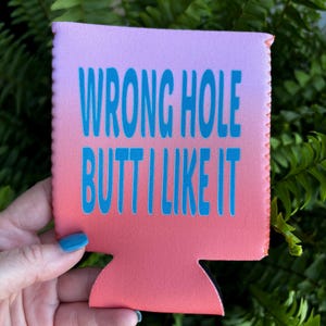 May include: A rectangular foam beverage insulator featuring a pink to orange gradient background. The text WRONG HOLE BUTT I LIKE IT is printed in a bold, bright blue sans-serif font across the center of the front surface of the sleeve.