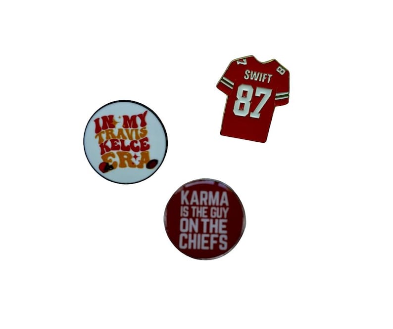 Puede incluir: Tres pines redondos con dise&ntilde;os rojos, blancos y dorados. El primer pin dice "In My Travis Kelce Era". El segundo pin dice "Karma Is The Guy On The Chiefs". El tercer pin es una camiseta de f&uacute;tbol roja con el n&uacute;mero 87 y el nombre "Swift" en la espalda.