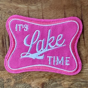 May include: A pink embroidered patch with the words "IT'S Lake TIME" in white script. The patch has a wavy, retro-style shape with a light pink border and a textured fabric appearance.