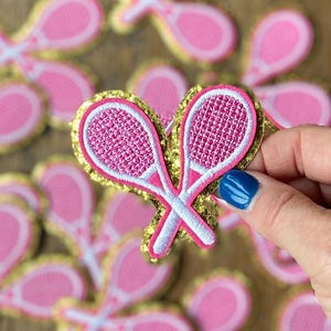 May include: Pink and white tennis racket shaped iron-on patches with gold glitter trim. The patches are arranged in a pile with one patch being held by a hand.