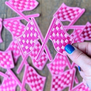 Op de afbeelding: Een stapel roze geruite racevlaggen met één vlag die door een hand omhoog wordt gehouden. De vlaggen zijn geborduurd met een geruit patroon.