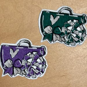 Op de afbeelding: Twee geborduurde patches in de vorm van een megafoon en pompons. Eén is paars met een wit hart en zilveren strik. De andere is groen met een wit hart en zilveren strik. Beide hebben een witte rand.