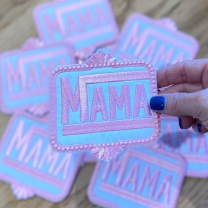 May include: A set of six pink and blue embroidered iron-on patches with the word "MAMA" in pink lettering.
