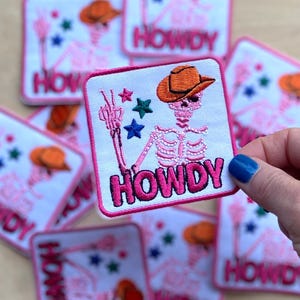 Könnte beinhalten: Ein Set aus rosa und weißen gestickten Bügeletiketten mit einem Skelett, das einen Cowboyhut trägt und "Howdy" sagt.