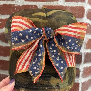Könnte beinhalten: Tarnfarbene Baseballkappe mit einer großen Schleife mit einem verwitterten amerikanischen Flaggenmuster. Die Schleife hat rot-weiße Streifen und blaue Abschnitte mit weißen Sternen. Die Kappe hat einen Mesh-Rücken und einen gebogenen Schirm.