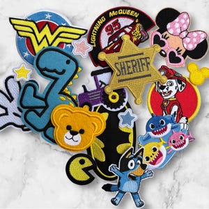 Könnte beinhalten: Eine Sammlung farbenfroher, gestickter Aufnäher mit Zeichentrickfiguren und Symbolen. Enthält ein Wonder Woman-Logo, einen Dinosaurier, einen Bären, ein Sheriff-Abzeichen, eine Minnie Mouse, einen Dalmatiner, eine Haifischfamilie und einen Bluey-Charakter.