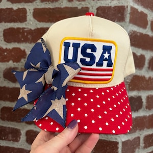May include: A baseball cap with a cream-coloured crown and a red brim dotted with white spots. The cap features a patch with the word "USA" in blue and red, and a blue bow with star patterns.