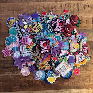 Puede incluir: Una colección de parches bordados coloridos con varios diseños, incluyendo estrellas, flores, corazones y texto. Los parches presentan una variedad de colores, incluyendo rosa, morado, azul y rojo. Algunos parches tienen texto como "Teach Love Inspire" y "Have a Nice Day".