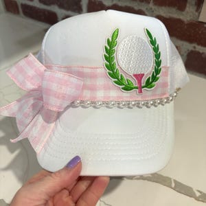 May include: White trucker hat with a golf ball and wreath design. The hat features a pink gingham ribbon, a pearl bead trim, and a large pink bow. The golf ball is white with a green wreath and pink tee.