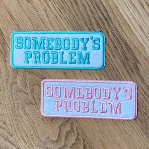 May include: Two white embroidered patches with the text "Somebody's Problem" in turquoise and pink lettering.