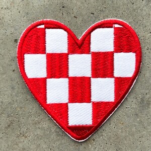 Puede incluir: Parche bordado en forma de corazón con un diseño a cuadros rojos y blancos. El parche tiene un borde rojo y probablemente está diseñado para coser o planchar sobre tela. El diseño es una forma de corazón clásica.