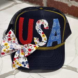 Pode incluir: Boné de caminhoneiro azul marinho com as letras "USA" aplicadas na frente. O "U" é vermelho com bolinhas brancas, o "S" é floral e o "A" é azul com um padrão paisley. Uma fita floral é anexada. O chapéu tem uma borda amarela.