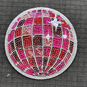 May include: Round, sequined patch featuring a globe design. The globe is made up of red, pink, and magenta sequins, outlined with silver glitter. The patch is on a dark gray fabric background.
