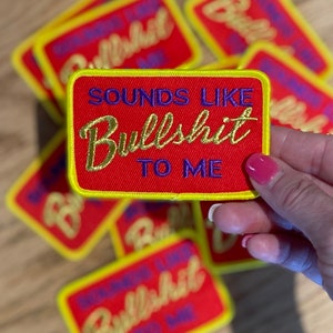 Puede incluir: Parche rojo bordado con el texto "SOUNDS LIKE BULLSHIT TO ME" en dorado y azul. El parche tiene un borde amarillo. Varios parches son visibles.