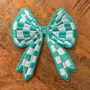 May include: A green and white checkered bow-shaped patch. The bow is made of fabric and has a stitched design.