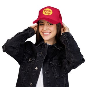 May include: A bright red trucker hat featuring a yellow oval patch on the front panel. The patch contains the text Kansas City written in a cursive red font. The hat has a structured front crown and a mesh back panel with an adjustable snap closure.