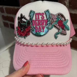 May include: A pink trucker hat with a white front panel. The hat features the text "IT'S DERBY DAY" in pink and teal sequins, along with horse head and horseshoe designs. A string of pearls adorns the brim.