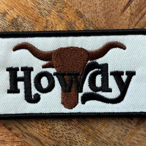 May include: White embroidered patch with a brown steer head and the word "Howdy" in black lettering.