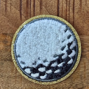 May include: Circular embroidered patch resembling a golf ball. The patch features a white, textured upper half and a gray, dimpled lower half, outlined with a gold glitter border. The design is set against a wooden surface.