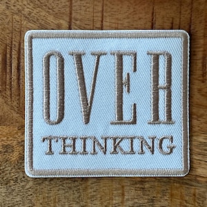 May include: Square embroidered patch with the words "OVER THINKING" in brown thread on a white background. The patch has a brown border and is set against a wooden surface.
