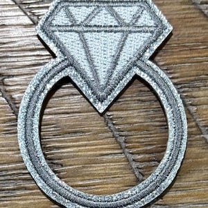 May include: A silver diamond ring patch with a glittery finish. The patch features a large, detailed diamond shape atop a circular band. The ring is embroidered with silver thread, creating a textured appearance. The patch is designed to be sewn or ironed onto fabric.
