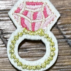 May include: A white felt patch in the shape of a diamond ring. The ring features a pink and white embroidered diamond and a gold embroidered band. The ring is a decorative item.