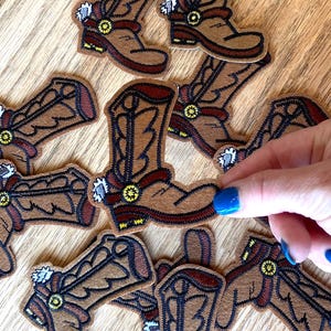 May include: Eight brown felt cowboy boot patches with black stitching and yellow accents. The boots have a spur on the heel and a buckle on the top of the boot.