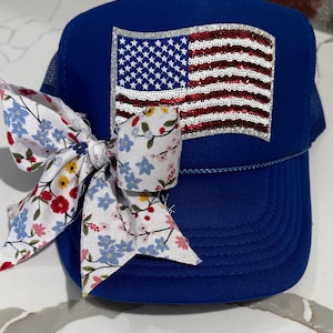 May include: A blue trucker hat with a sequined American flag patch. A floral bow in red, white, and blue is attached to the side of the hat. The hat has a mesh back.