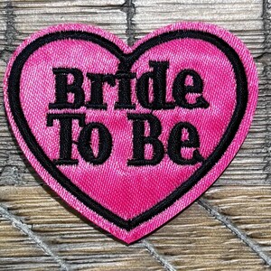 May include: A pink heart-shaped patch with the words "Bride To Be" embroidered in black. The patch has a black border and is likely for a bridal shower or bachelorette party.