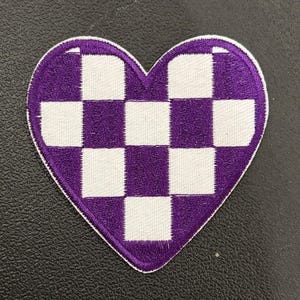 May include: A heart-shaped patch with a purple and white checkered pattern. The patch has a white border and is made of fabric. The design is a classic checkerboard, with alternating squares of purple and white.