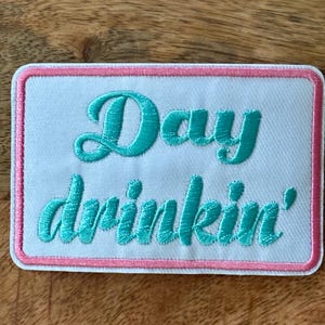 Puede incluir: Parche bordado con las palabras "Day drinkin'" en guión turquesa sobre un fondo blanco. El parche tiene un borde rosa y una forma rectangular. Para chaquetas o bolsos.