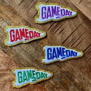 May include: Four white felt pennant patches with gold glitter trim. Each patch has the word "GAMEDAY" embroidered in a different color: red, purple, blue, and green.